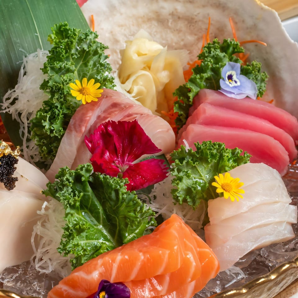 Assorted sashimi platter with tuna, salmon, and white fish, garnished with kale, flowers, and ginger.
