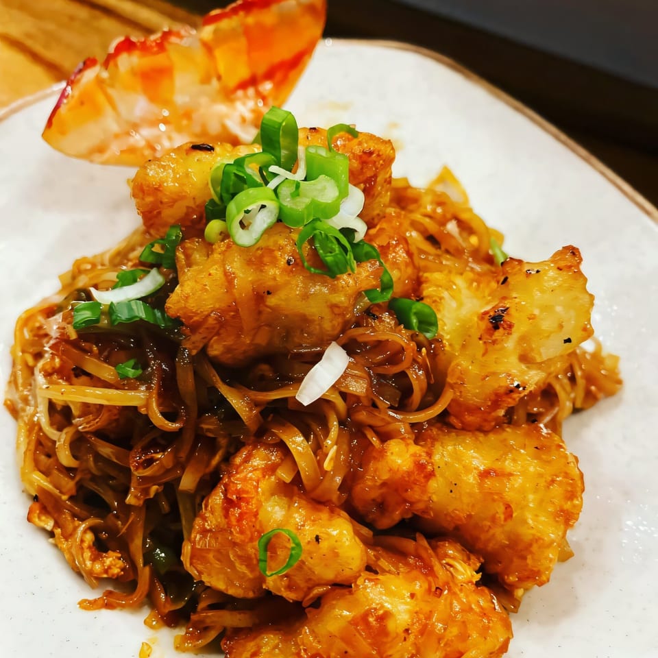 Thai noodles with crispy fried chicken pieces, garnished with fresh green onions and a lobster tail.