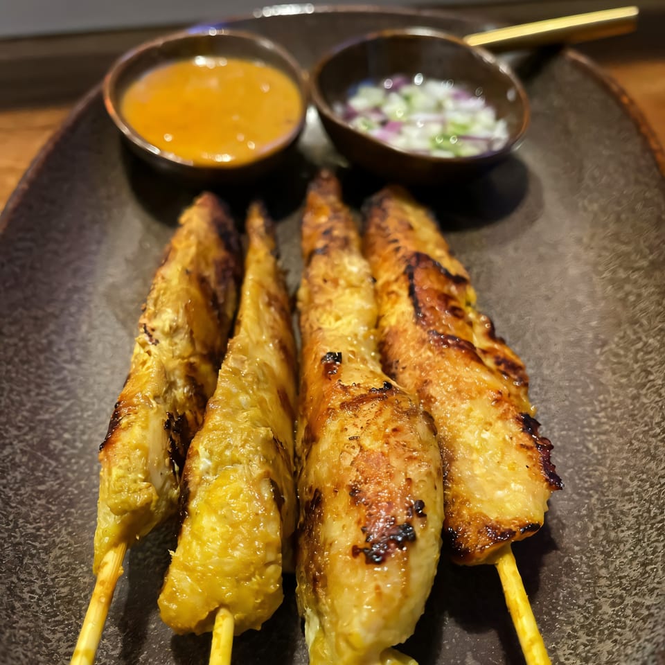 Four grilled chicken satay skewers with two dipping sauces on a dark plate.